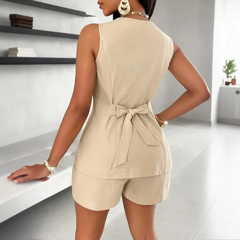 A woman stands with her back to the camera, wearing a beige sleeveless vest with a tie detail at the waist and matching shorts. The background features a modern, well-lit room with minimalistic decor and a large window.